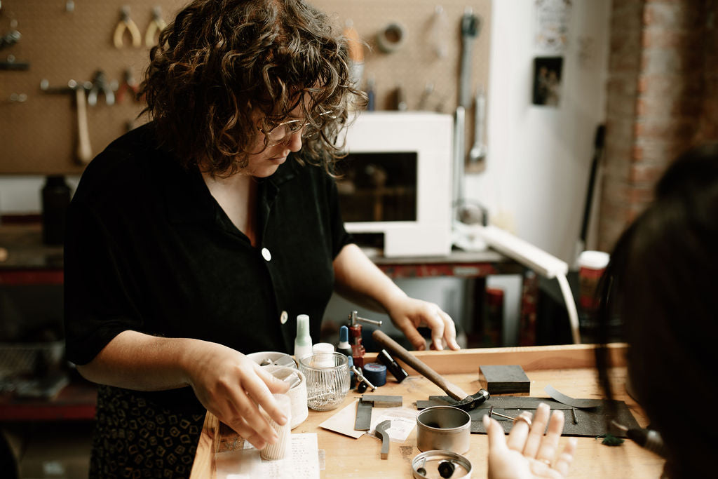 woman looking down at jeweller’s bench full of tools with person sitting holding a ring up