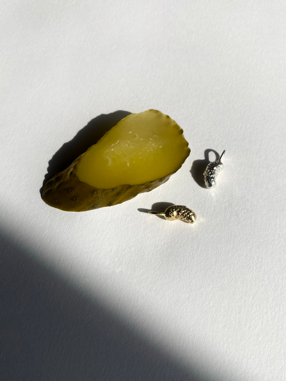 Silver and gold pickle charms beside pickle slice on white background