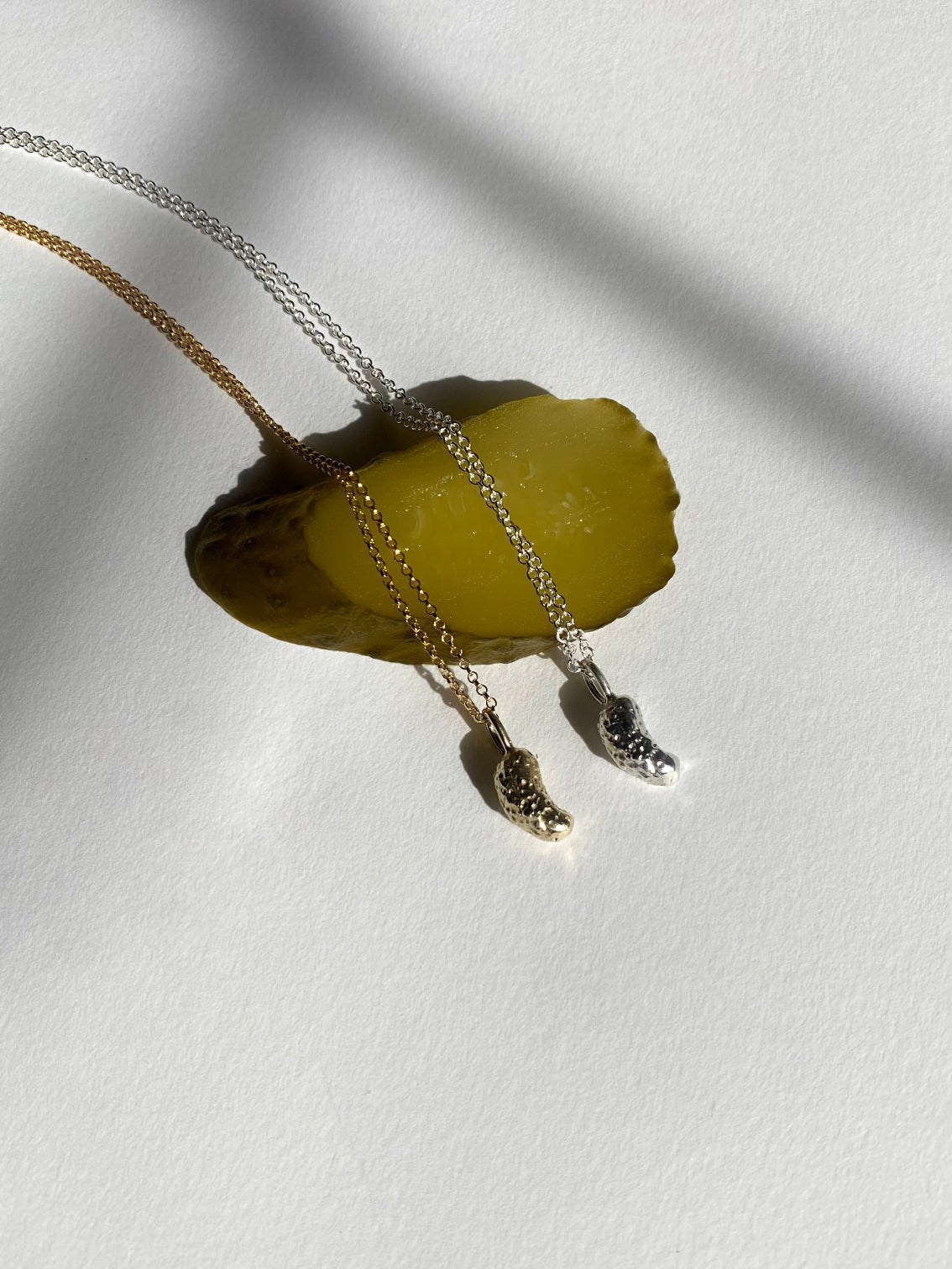 Silver and gold pickle charm necklaces resting on pickle slice on white background
