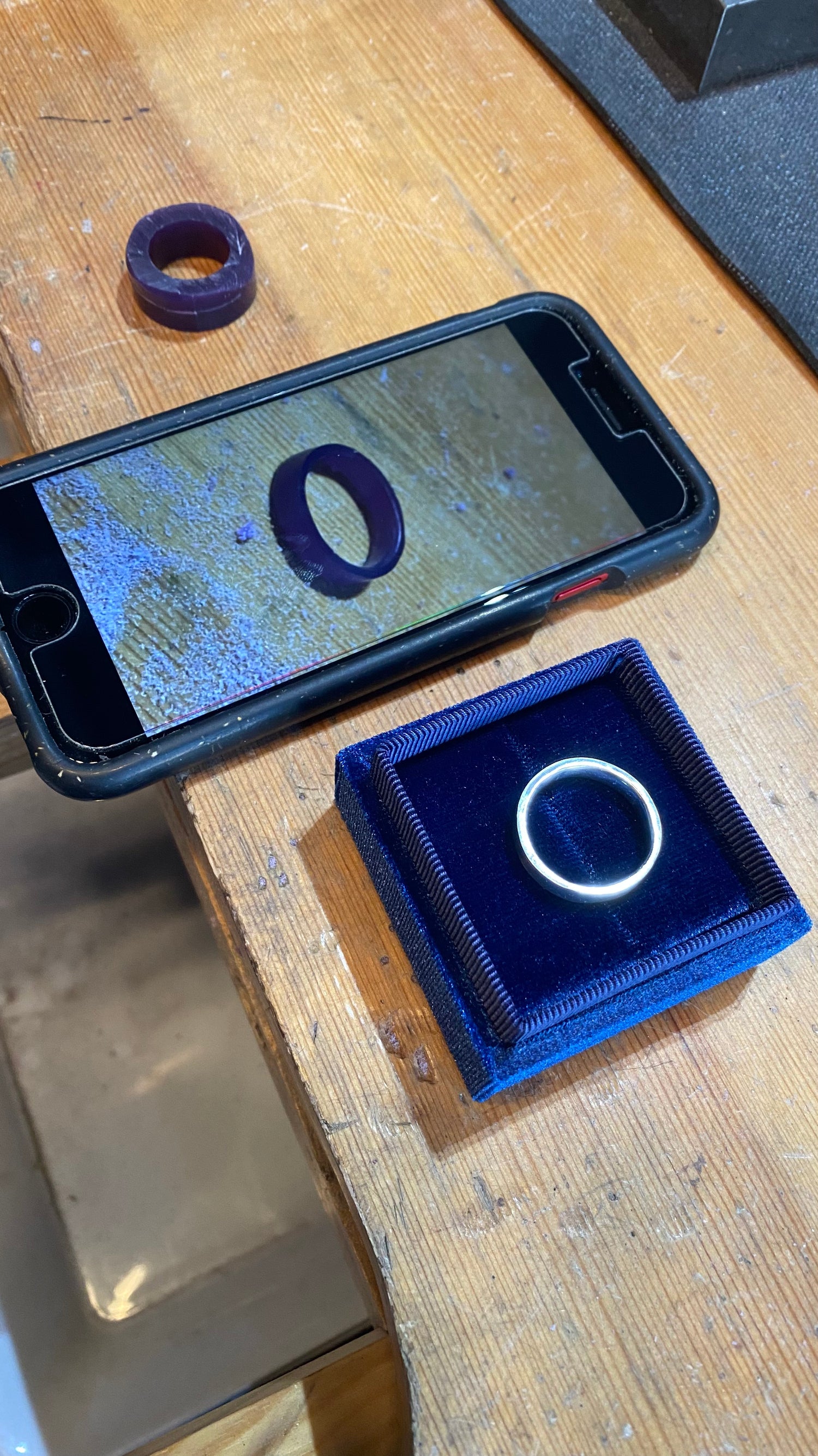 Layout on wood table of wax ring tube, phone with carved wax ring, and final white gold ring in blue velvet box