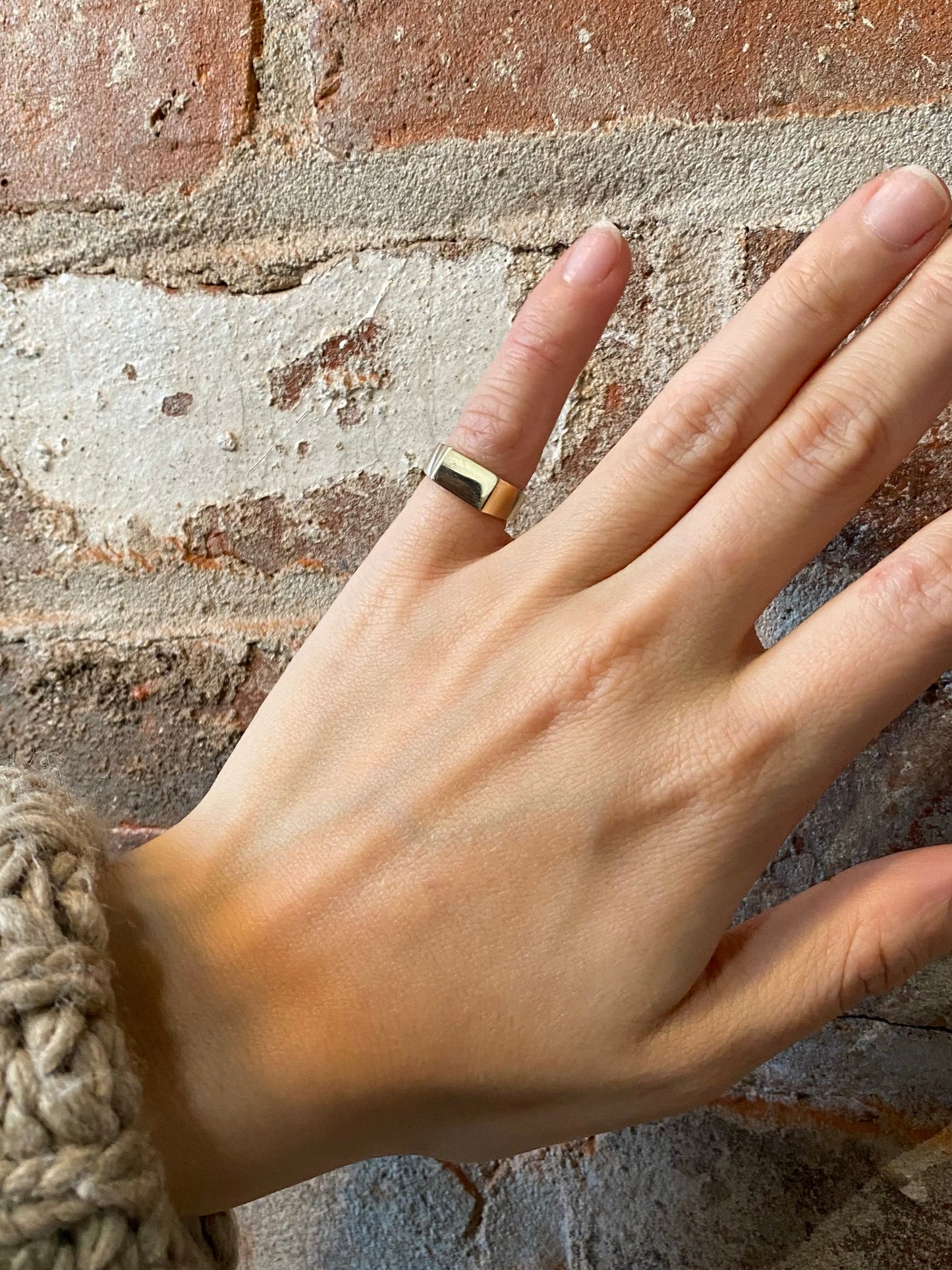 Square gold pinky ring on hand in front of brick wall